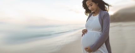 A pregnant person stands on a beach, cradling their belly. 