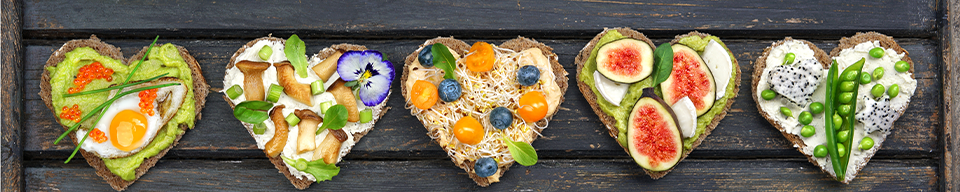 five heart-shaped pieces of toast sit side-by-side topped with various fruits, veggies, and spreads