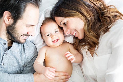 A man, a baby, and a woman lay together, all smiling. The man and woman llok at the baby while the baby looks at the camera.