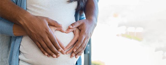 Two sets of hands form a heart shape over a pregnant belly.