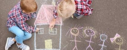 Two children draw a house and a stick figure family with sidewalk chalk.