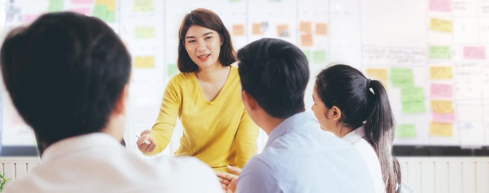 A woman speaks with group of people in front of a board with several colored notes pinned to it.